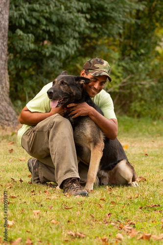 A man kneels down and hugs his german shepherd dog on a grassy lawn with trees in the background. 