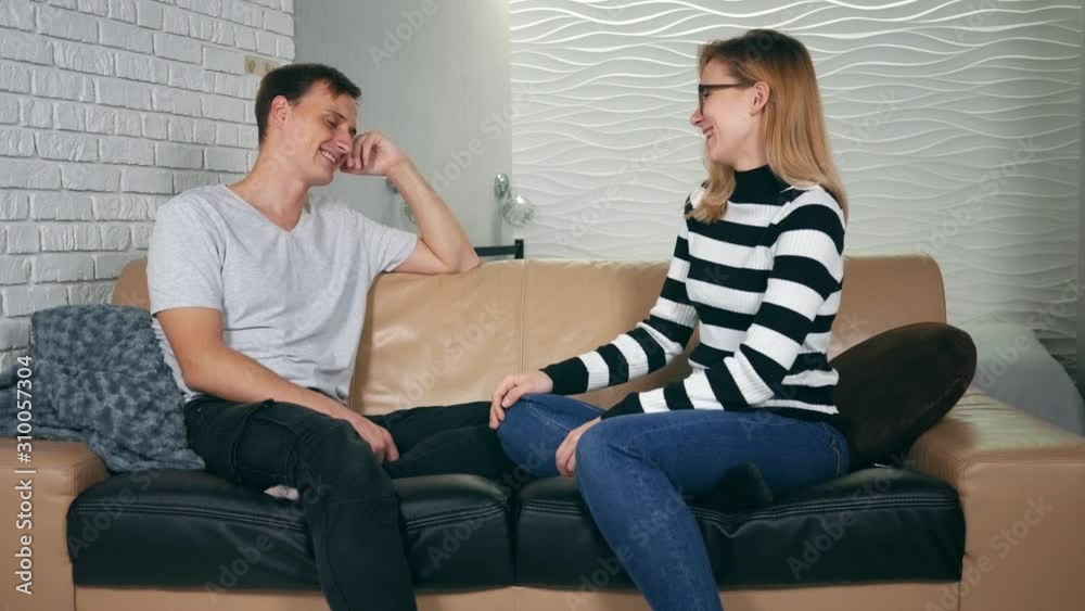 Happy young couple sitting on couch and talking and laughing at home. Communication, relationships and people concept - happy couple sitting on sofa and talking at home Happy young couple sitting on couch and talking and laughing at home. Communication, relationships and people concept - happy couple sitting on sofa and talking at home