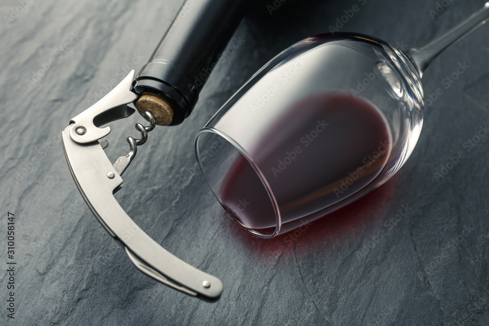 Stainless corkscrew in a cork of bottle neck and wine glass with wine lying on a black slate background