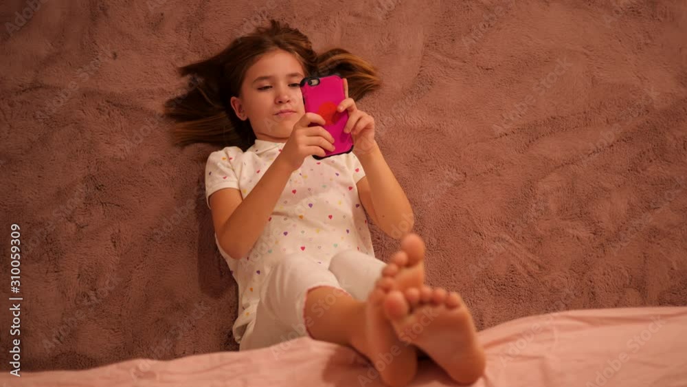 Kid girl of school age, lie on a floor in bedroom texting messages to friends using smartphone having fun before go to bed