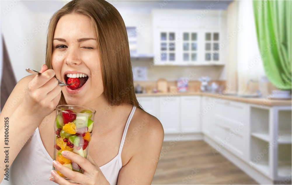 Young woman eating fruits from glass on blurred kitchen background Young woman eating fruits from glass on blurred kitchen background