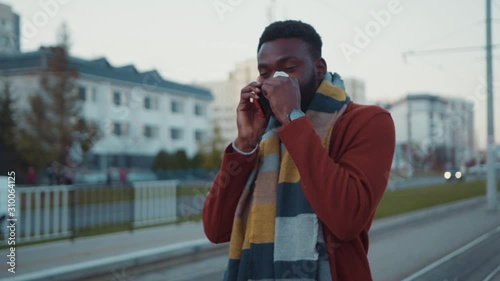 Sick unwell handsome black man in warm coat talking on mobile phone sneezing allergy with papertissue waiting for public transport at tram stop. photo