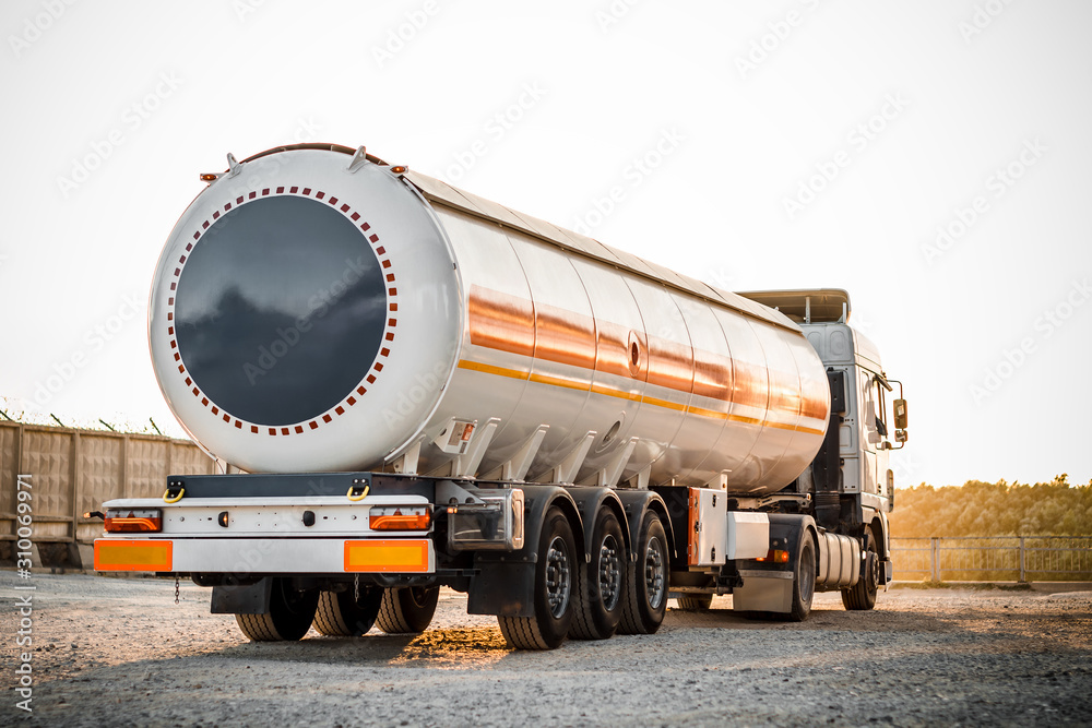 Truck with trailer, tank with flammable liquid, under a large bridge at the pier on the river bank, sunset light, white and red cars standing on gravel