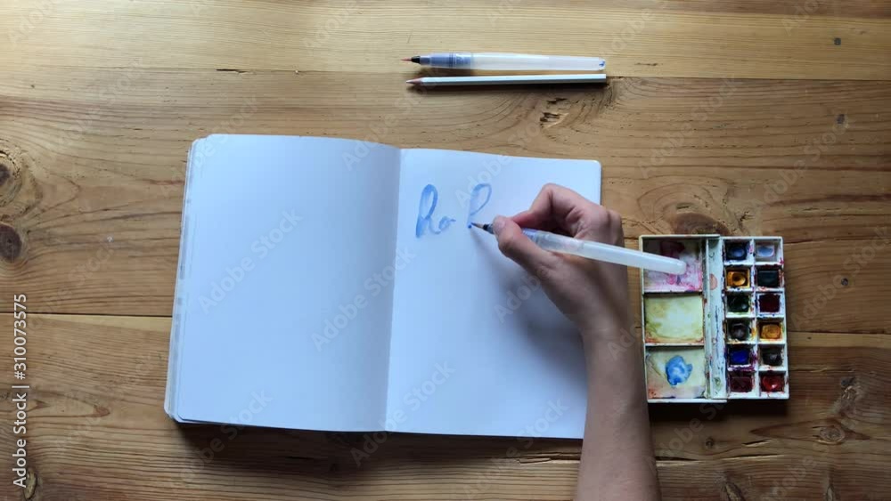 Young Woman artist draw with watercolor paints with a brush in a bullet journal pages. Blue watercolor painter on white canvas. Woman hands painting a christmas message. Slow motion.