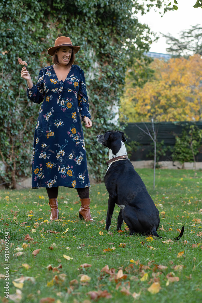 Portrait of young beautiful woman  in brown hat and blue dress playing with greyhound dog on the grass with leaves