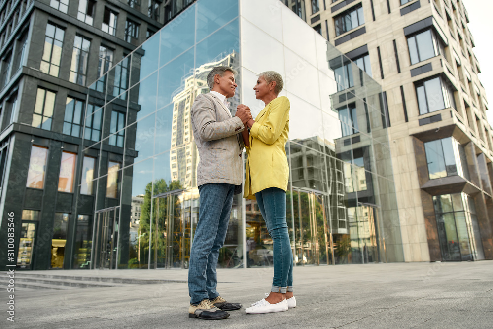 I wish to be with you forever. Full-length of elegant and stylish mature couple holding hands and looking at each other while standing against modern glass building