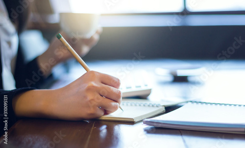 Close-up young businesswoman writing notebook and holding coffee cup to drink while she working on desk