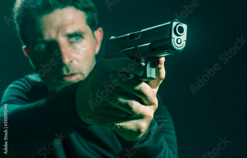 shallow depth of focus action portrait of serious and attractive hitman or special agent man holding gun pointing the weapon isolated on dark background