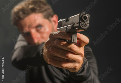 shallow depth of focus action portrait of serious and attractive hitman or special agent man holding gun pointing the weapon isolated on dark background