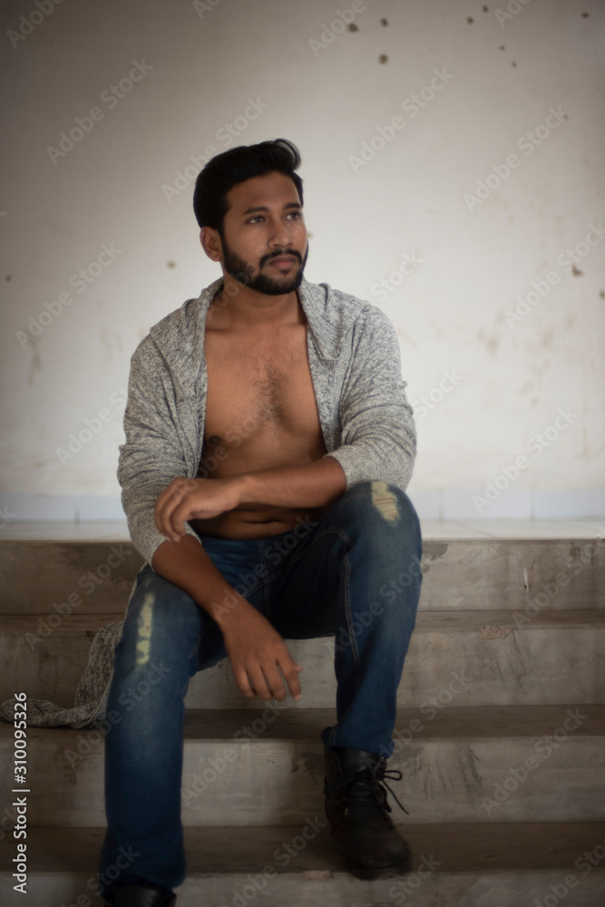 An young tall, dark and handsome Indian Bengali man in a front open western jacket and jeans sitting on a stair case in white background. Indian lifestyle and fashion. An young tall, dark and handsome Indian Bengali man in a front open western jacket and jeans sitting on a stair case in white background. Indian lifestyle and fashion.