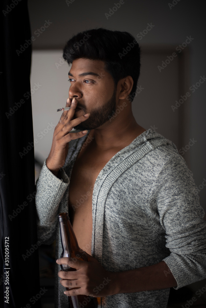An young tall, dark and handsome Indian Bengali man in a front open western jacket smoking while looking outside standing in front of a window in studio background. Indian lifestyle and fashion. An young tall, dark and handsome Indian Bengali man in a front open western jacket smoking while looking outside standing in front of a window in studio background. Indian lifestyle and fashion.