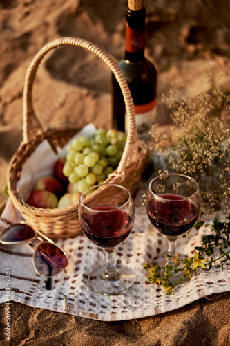 On the sand lies a blanket on which are glasses with red wine, a bottle of wine and a knitted fruit basket. Romantic dinner by the sea for two.