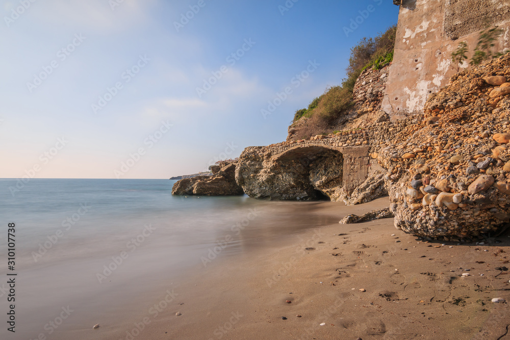 Beach section on the Mediterranean coast in Nerja. Sandy beach on the Spanish coast of Costa del Sol with rocks and stone arch on sunny day with blue sky and clouds