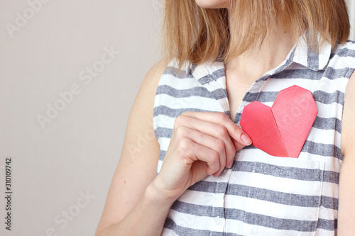 caucasian woman holding origami red paper heart, puts valentine in breast pocket. Insurance, charity donation concept. Place for text, copy space, selective focus