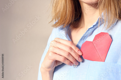 caucasian woman holding origami red paper heart, puts valentine in breast pocket. Insurance, charity donation concept. Place for text, copy space, selective focus