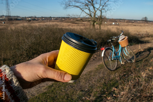 In hand a yellow paper corrugated cup with coffee on a background of nature and a bicycle photo