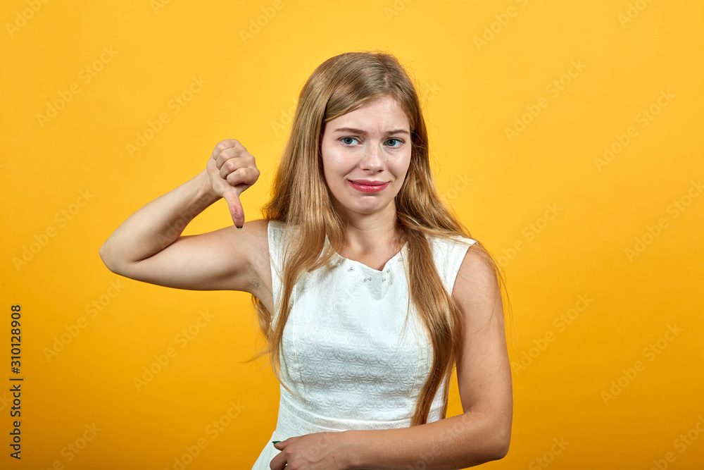 Charming blonde young caucasian woman pointing thumb down, looking disappointed over isolated orange background wearing white shirt. Lifestyle concept Charming blonde young caucasian woman pointing thumb down, looking disappointed over isolated orange background wearing white shirt. Lifestyle concept