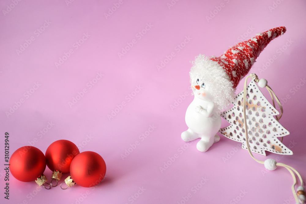 Obraz premium Funny cute snowman in a red hat near the Christmas tree and three red Christmas balls on a pink background with copy space. Christmas and New Year concept, minimalism.