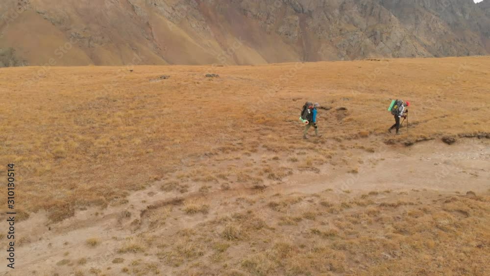 Aerial view of a couple of travelers man and woman with large backpacks in hats and sunglasses walk along an alpine plateau surrounded by epic mountains Aerial view of a couple of travelers man and woman with large backpacks in hats and sunglasses walk along an alpine plateau surrounded by epic mountains