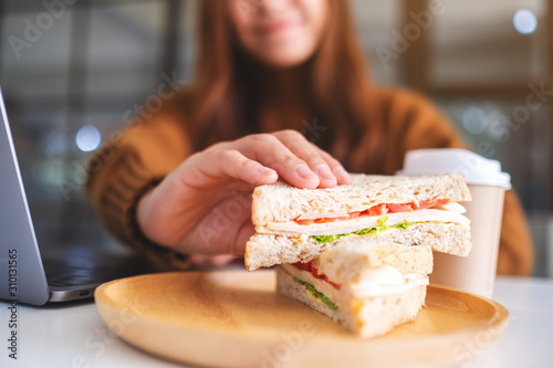 An asian woman holding and eating whole wheat sandwich while working on laptop computer photo