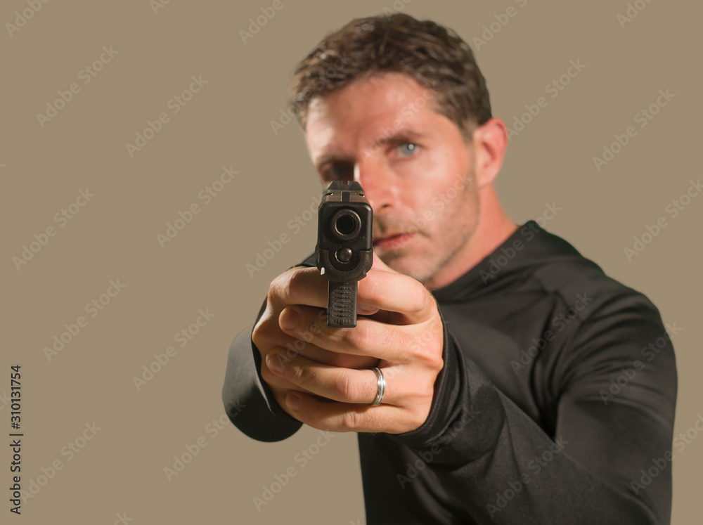 shallow depth of focus action portrait of serious and attractive hitman or special agent man holding gun pointing the handgun to the camera isolated on dark background shallow depth of focus action portrait of serious and attractive hitman or special agent man holding gun pointing the handgun to the camera isolated on dark background