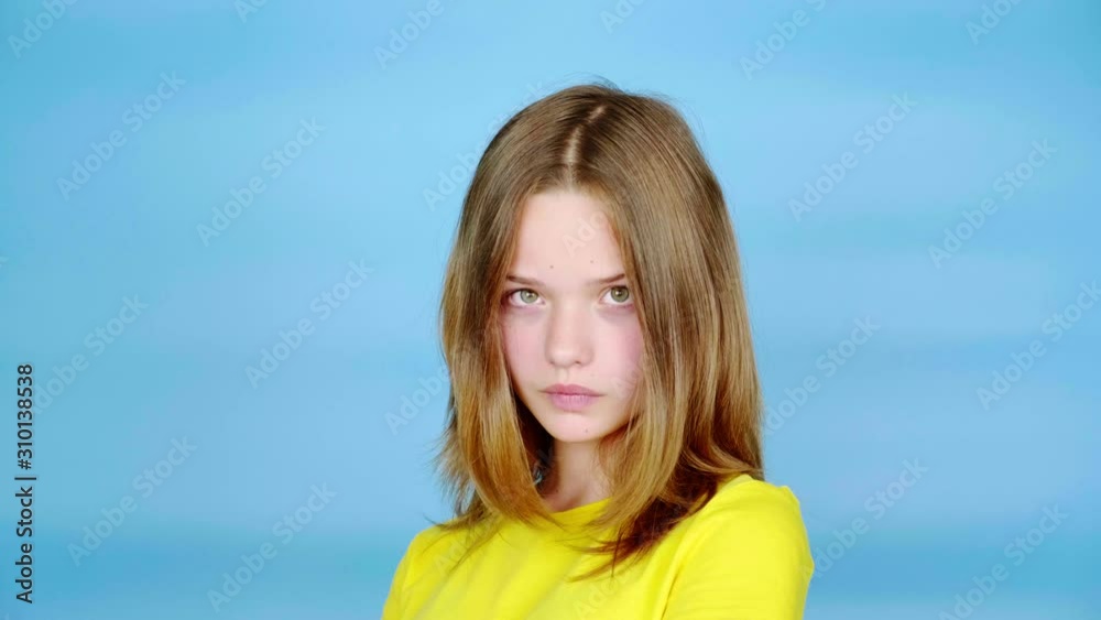 Teen girl in a yellow t-shirt is looking at the camera and shakes her head. Blue background with copy space. Teenager emotions. 4k footage