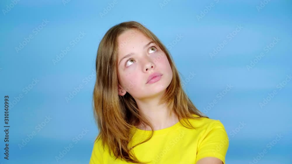 Teen girl in a yellow t-shirt is looking away and moves her hand. Blue background with copy space. Teenager emotions. 4k footage Teen girl in a yellow t-shirt is looking away and moves her hand. Blue background with copy space. Teenager emotions. 4k footage