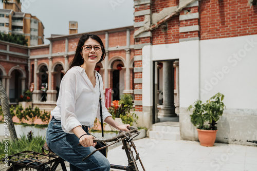 Happy young asian korean woman tourist on bike wearing white shirt and glasses smiling on sunny day. elegant lady traveler riding bicycle in chinese traditional building monument in spring time photo