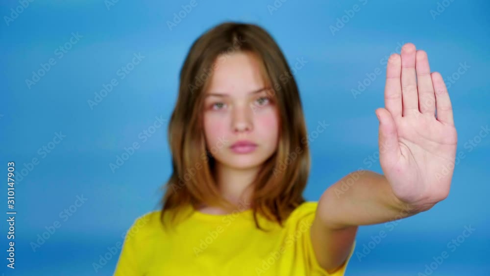 Teen girl in a yellow t-shirt is looks at camera, holds out her hand in a front and says stop. Blue background with copy space. Focused on the hand. Teenager emotions. 4k footage
