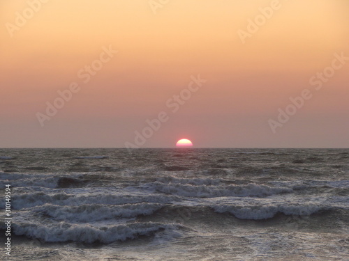 Fototapeta Naklejka Na Ścianę i Meble -  The stormy sea pushes a bright solar disk to the surface against a red-orange sky.