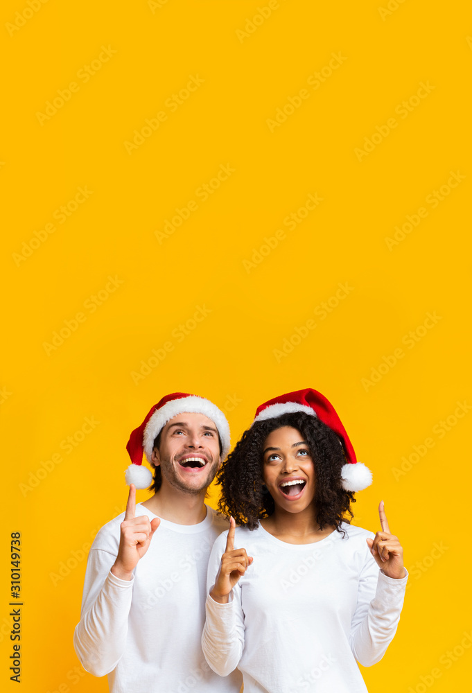 Multiracial couple wearing santa hats pointing upwards at copy space Multiracial couple wearing santa hats pointing upwards at copy space