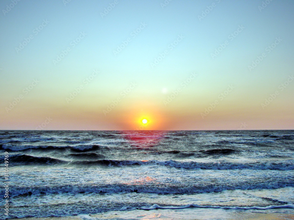 Fototapeta premium The stormy sea pushes a bright solar disk to the surface against a red-orange sky.