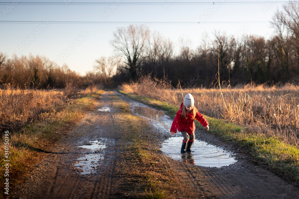 3 years old child jumping in a puddle in a country landscape ,with red jacket and wellies on a sunny winter day. Golden hour light.