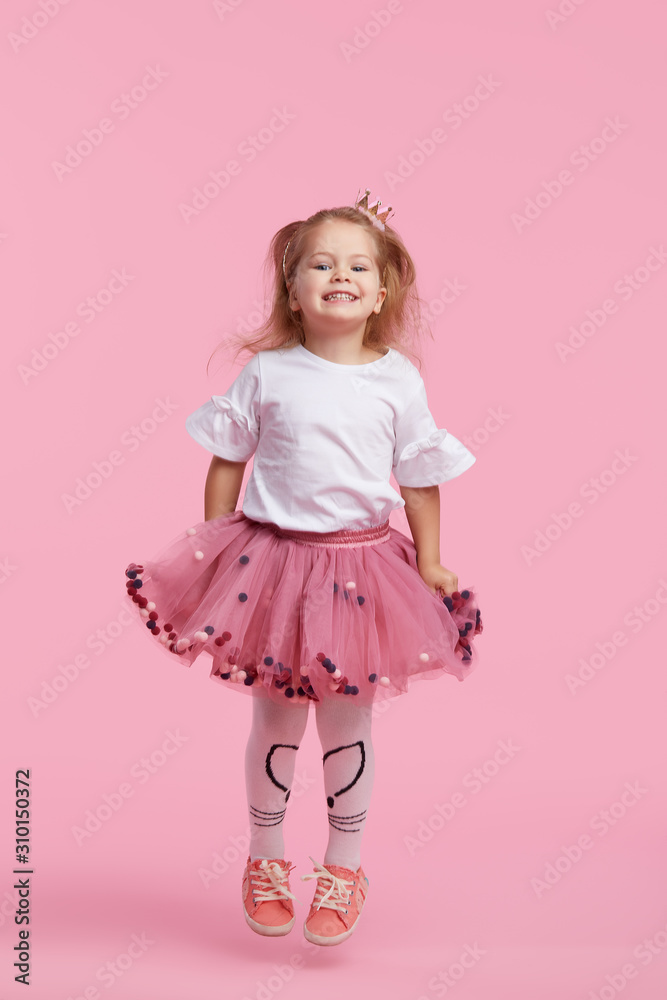 A cheerful little girl with in a tulle skirt and princess crown on her head isolated on a pink background. Celebrating a vibrant carnival for kids, birthday party, having fun