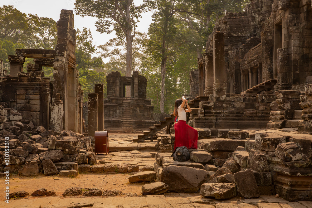 young female tourist in red dress taking a picture of the historic Bayon Temple, Angkor Wat, Cambodia