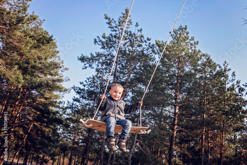 Smiling handsome blond boy in grey coat flying on a swing against blue sky