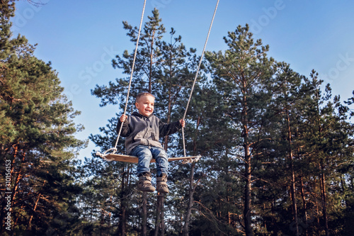 Smiling handsome blond boy in grey coat flying on a swing against blue sky