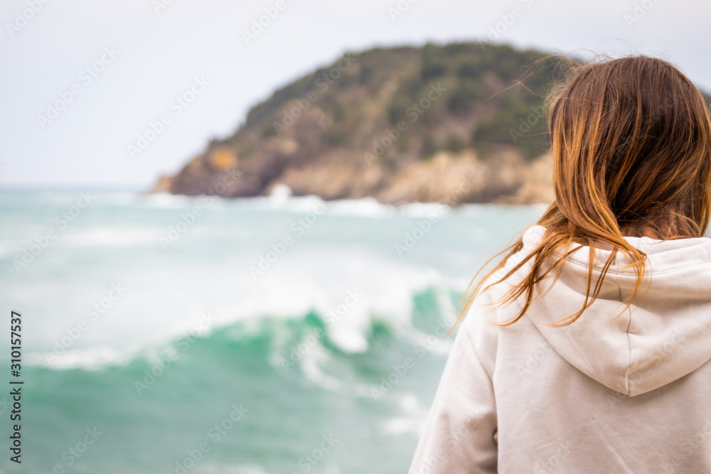 Young woman with sweatshirt sitting in front of the sea looking towards the horizon. There is a mountain in the background while breaking a wave in front of the woman.