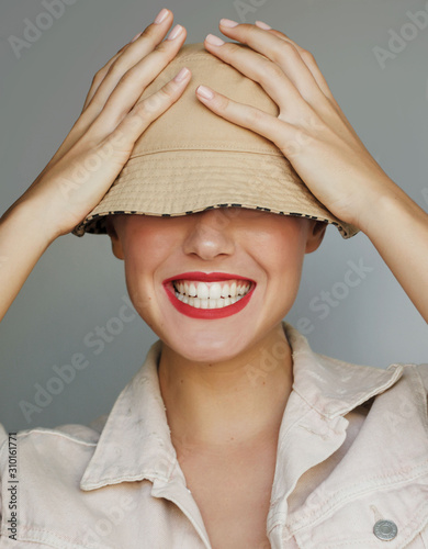 happy girl covering eyes with her hat laughing. Women laughing playfully wearing red lipsitck. Female covering her eyes with a bucket hat laughing. Woman with white teeth laughing.