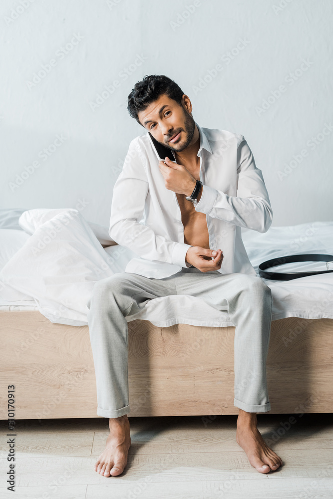 handsome bi-racial man talking on smartphone and sitting on bed in morning