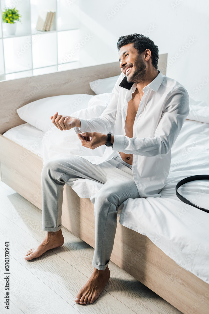 smiling and handsome bi-racial man talking on smartphone and sitting on bed in morning