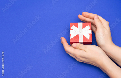 Female hands are holding a red gift box with a thin white ribbon as a present for Christmas, New Year, Mother's Day or anniversary on a blue table background with a copy space