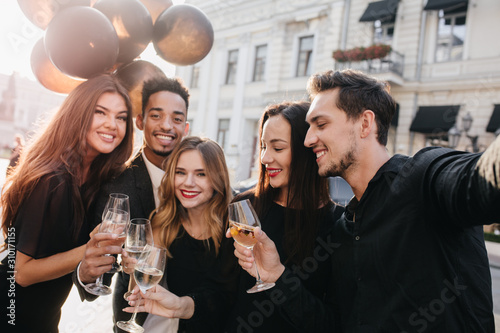 Blissful african man holding bunch of balloons and glass of wine in front of old building. Outdoor portrait of excited friends enjoying champagne during meeting after workday.
