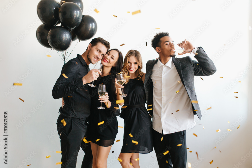 Indoor portrait of romantic pale girl with shiny curls enjoying wine with friends. Ladies in black dresses having fun at party with guys and dancing under confetti. Indoor portrait of romantic pale girl with shiny curls enjoying wine with friends. Ladies in black dresses having fun at party with guys and dancing under confetti.