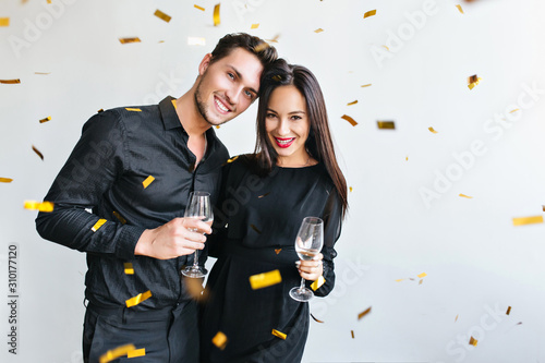 Happy woman with dark straight hair celebrating anniversary with husband. Indoor portrait of cheerful man with glass of wine gently embracing cute girlfriend in trendy dress.