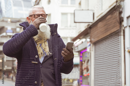 Handsome mature man drinking coffee in the morning near the job. Pensioner looking the smartphone and drinking hot tea. Man walking for the streets of London city - Image