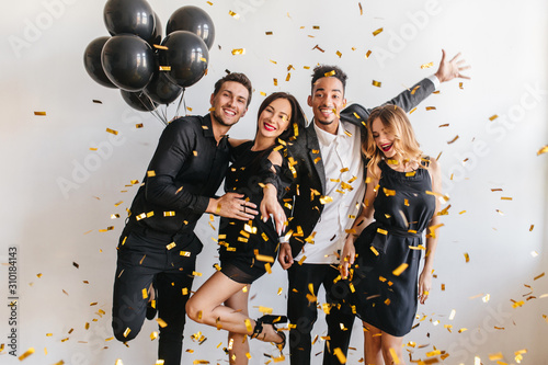 Curly blonde girl with red lips posing with eyes closed near excited african man dancing at party. Blissful brunette woman with balloons celebrating birthday with friends.