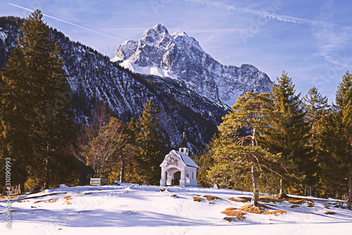 Kapelle auf Berg vor imposanter Hochgebirgskulisse im Schnee umgeben von Bergwald