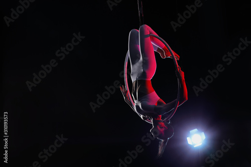 Young woman performing acrobatic element on aerial ring against dark background. Space for text photo