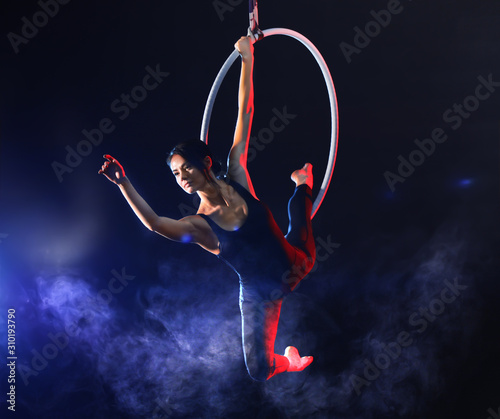 Young woman performing acrobatic element on aerial ring against dark background